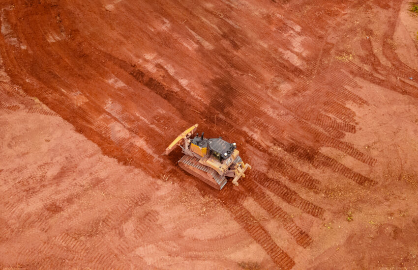 Bulldozer grading red dirt, tracks visible, sparse edge vegetation.