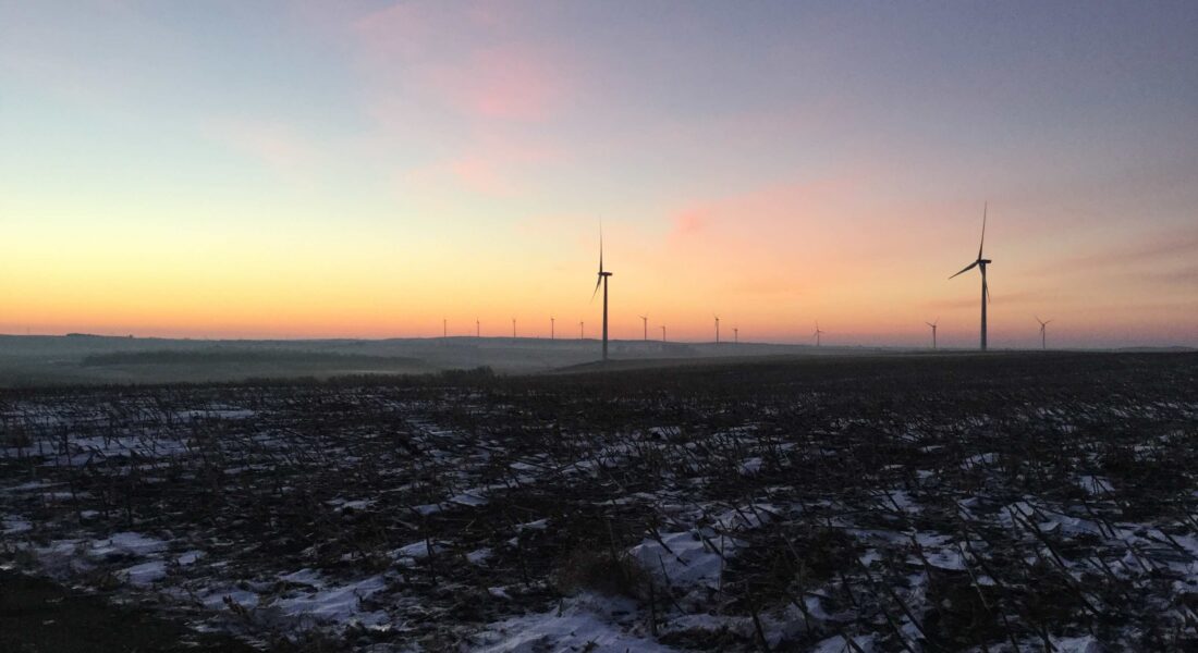 Snowy field with distant wind turbines beneath a pastel sunset.
