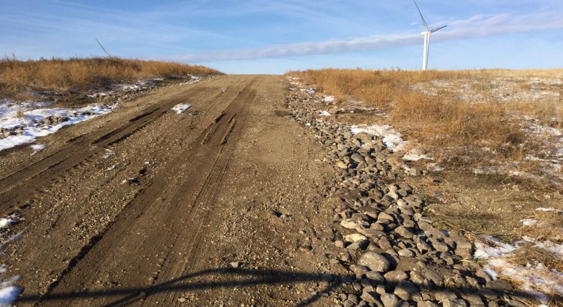 Uphill dirt road, patches of snow, wind turbine, blue sky.