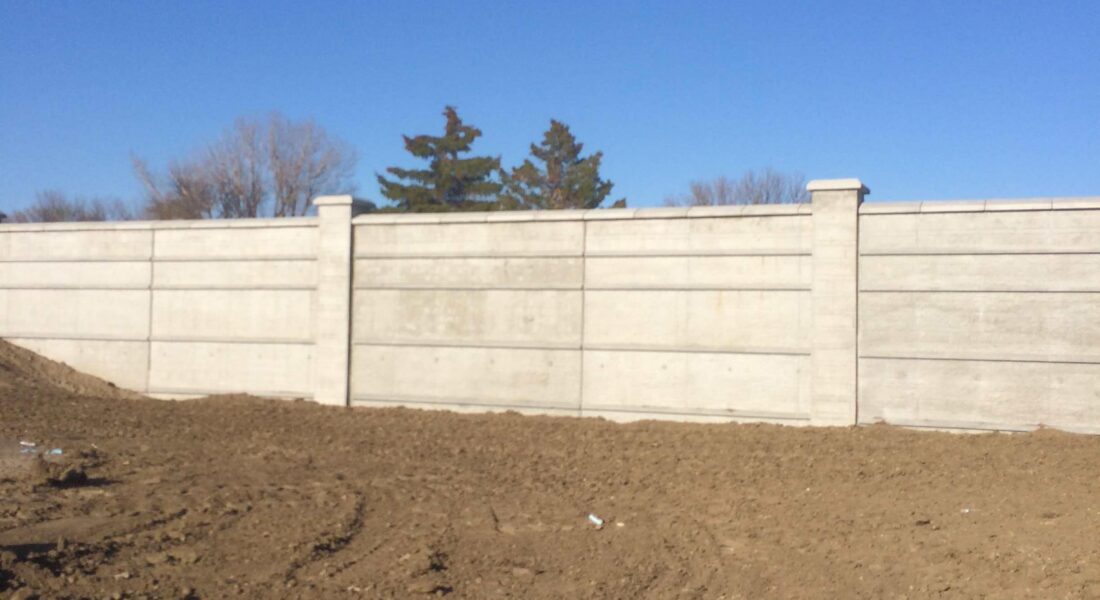 Dirt lot before tall concrete wall, three trees and blue sky.