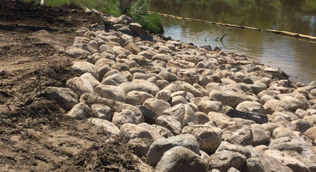 Excavator sets rocks by riverbank on a sunny day.