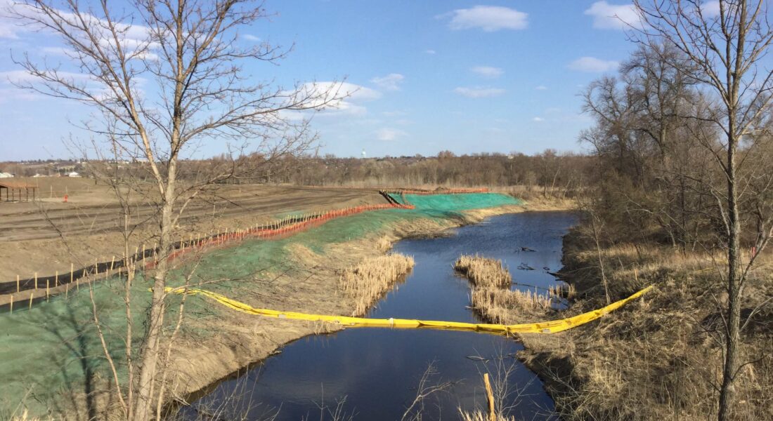 Stream with grassy banks, bare trees, erosion mats, yellow barrier.