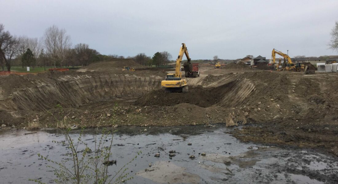 Three excavators work in a muddy depression under an overcast sky.