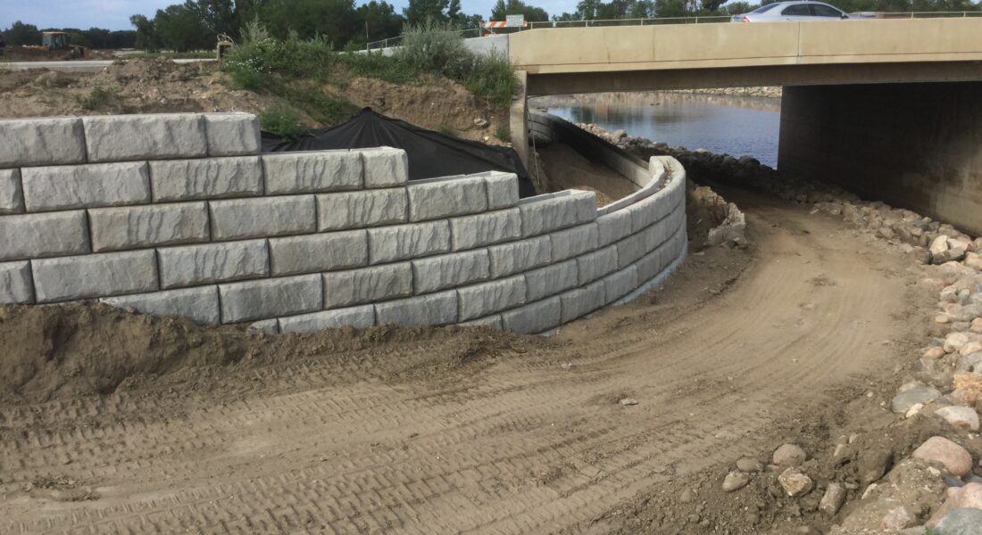 Curved retaining wall and dirt path under bridge, tire tracks.