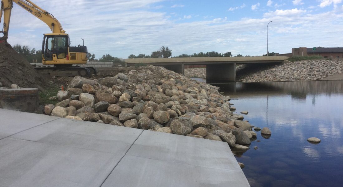 Excavator on rocks near water, bridge and clouds behind.