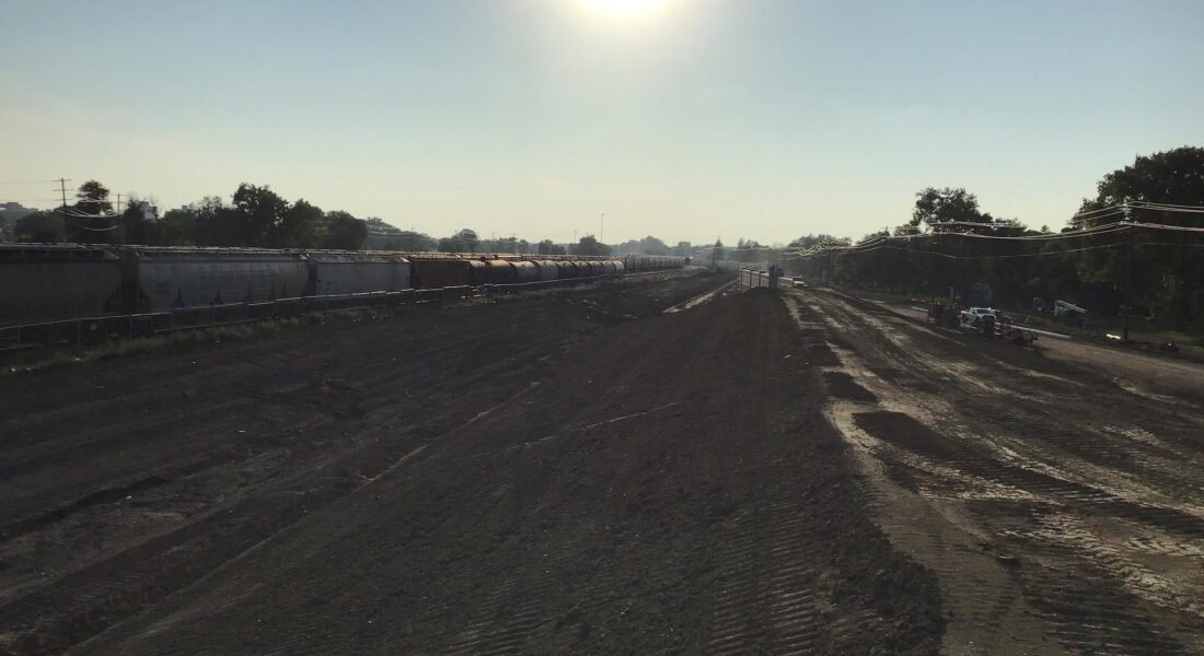 Sunny construction site, dirt ground, train cars left, trees behind.