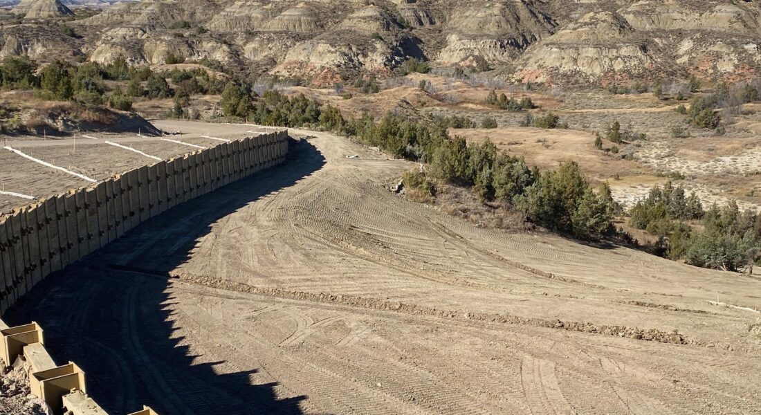 Curved concrete barrier on dry dirt path amid rocky hills.
