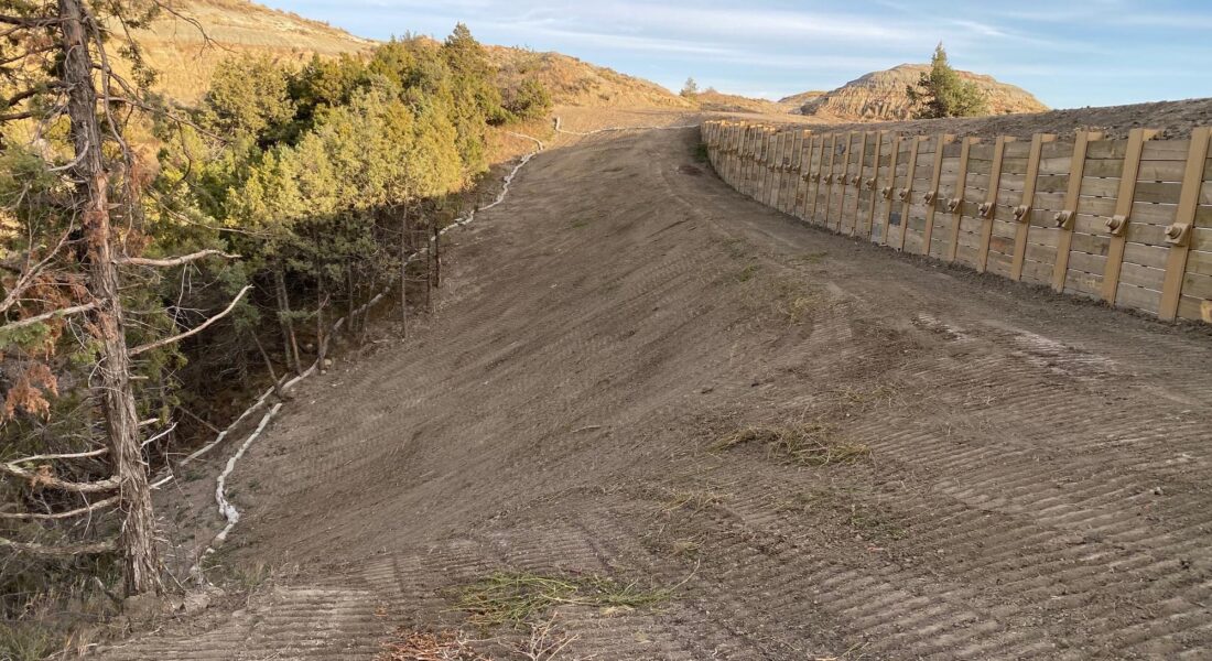 Dirt slope, tire tracks, wooden wall right, trees left, clear sky.