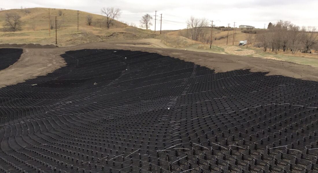 Black geogrid covers sloped site; bare trees and hills behind.