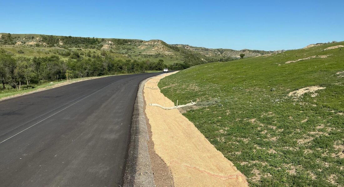Curved new road in grassy hills, erosion matting, yellow text overlay.