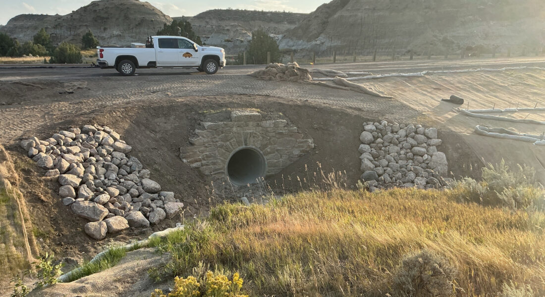 White pickup by culvert on gravel road, hilly grassy area, overlays present.