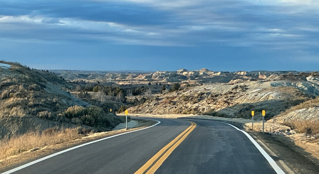 Curving highway crosses barren hills beneath partly cloudy sky.
