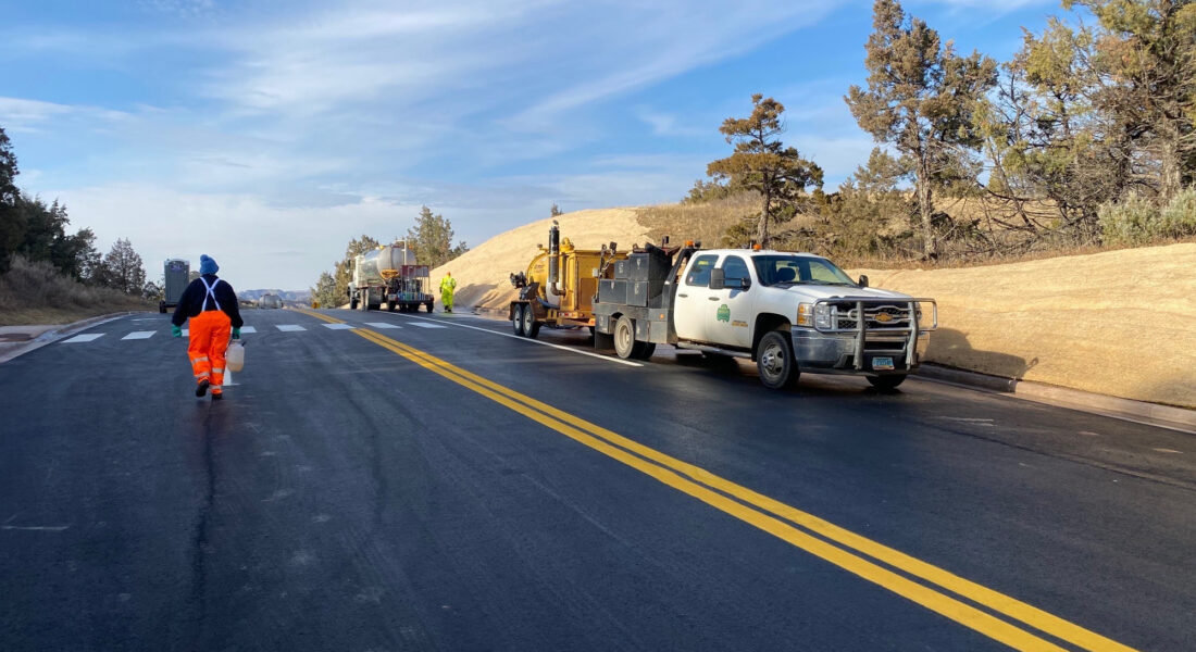 Workers and vehicles paint road lines on sunny rural morning.