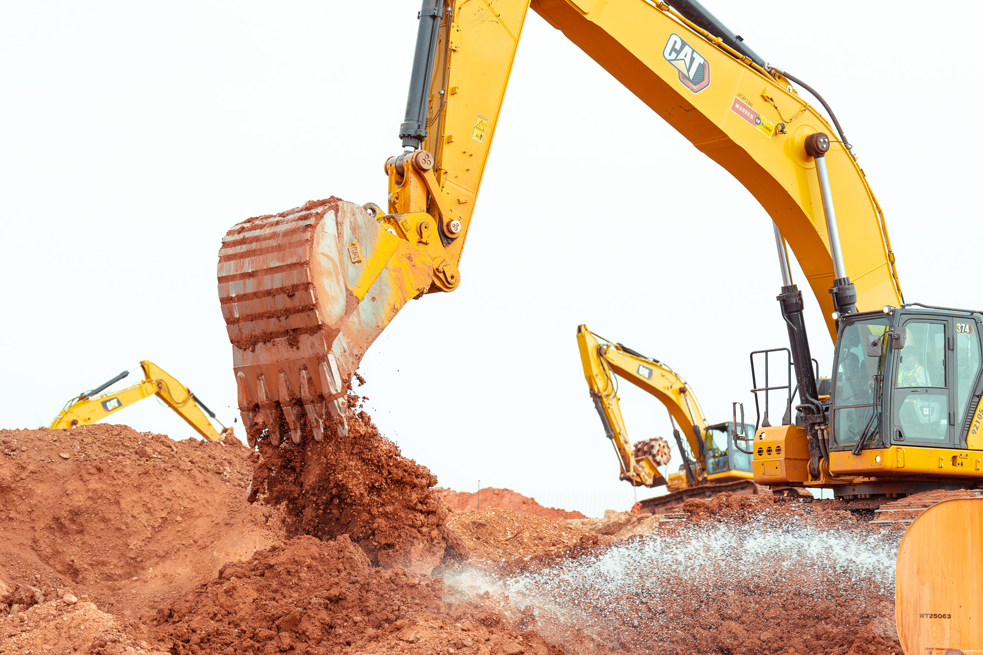Yellow excavator moves soil, spraying water; two others in back.