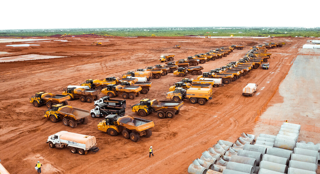 Construction vehicles and workers on red soil site, pipes nearby.