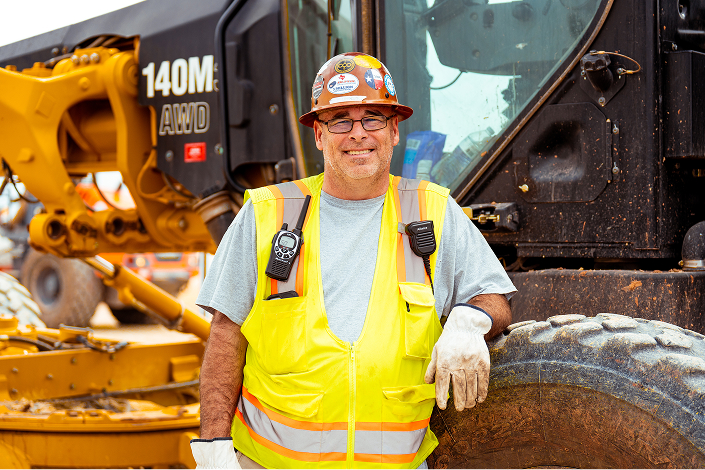 Smiling construction worker in safety gear beside heavy machinery.