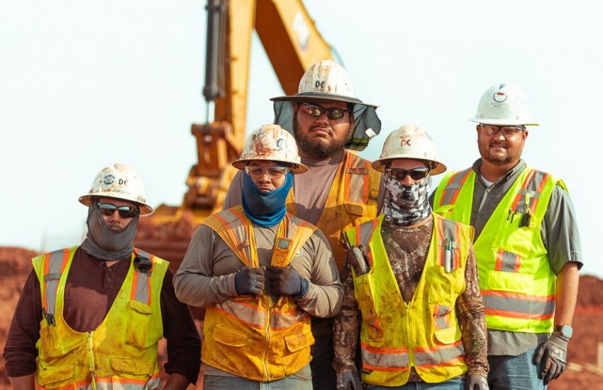 Five construction workers pose together in safety gear by machinery.