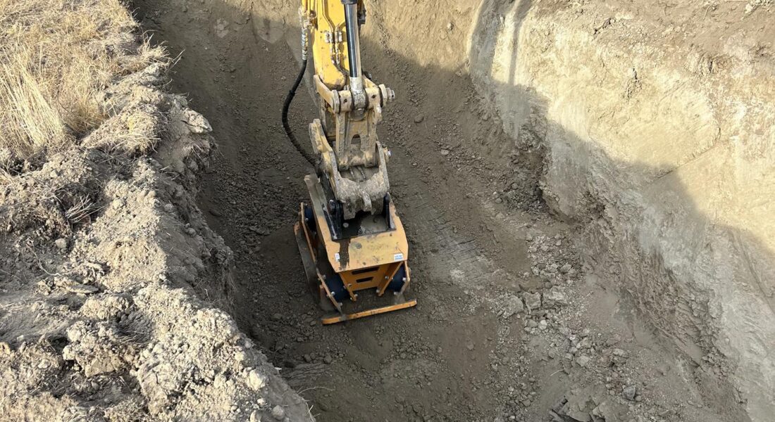 Yellow excavator compacts soil in deep construction trench.