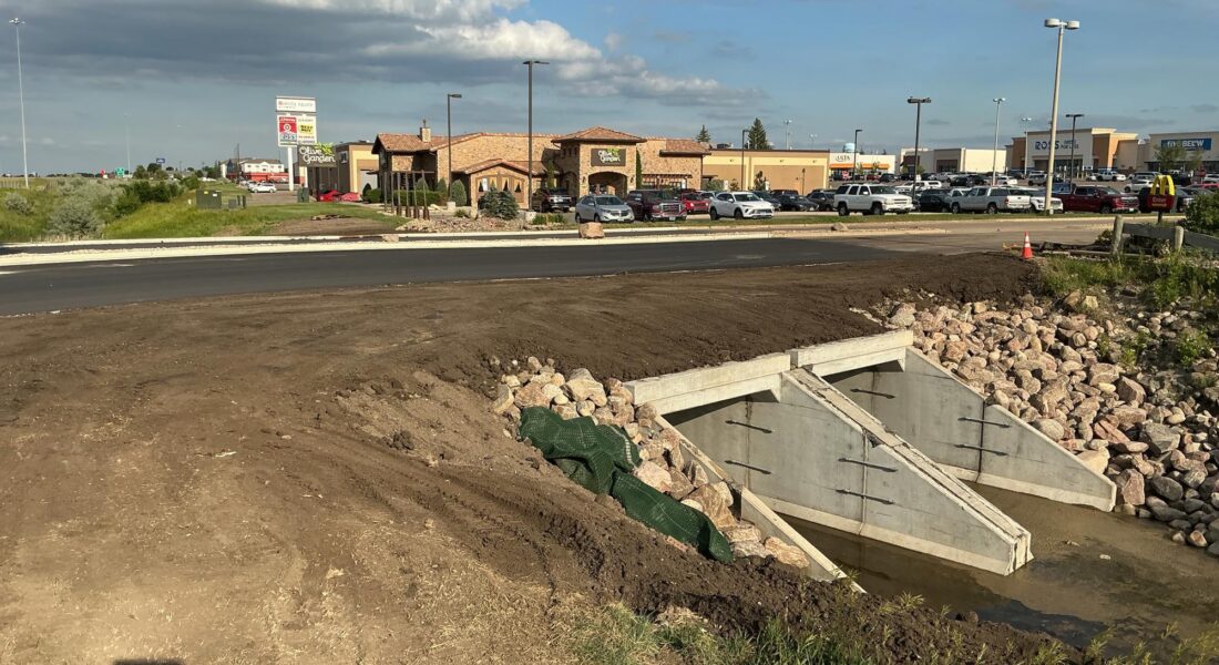 Concrete culvert with rock lining beneath road near buildings.