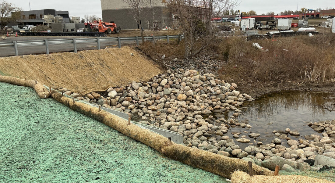 Rocky stormwater drainage by culvert, barriers, buildings behind.
