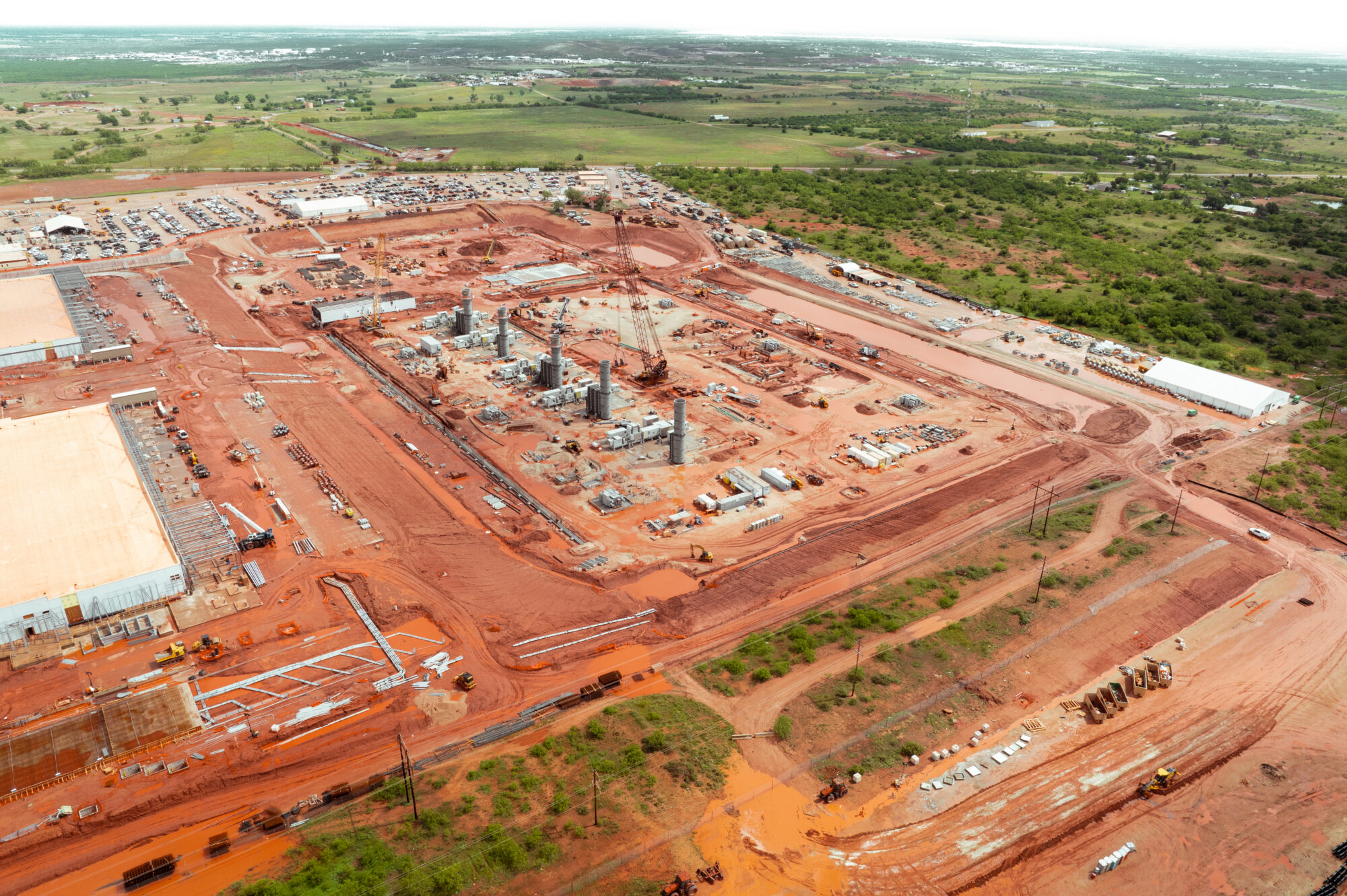 Aerial view: construction site, buildings, cranes, reddish soil, green fields.